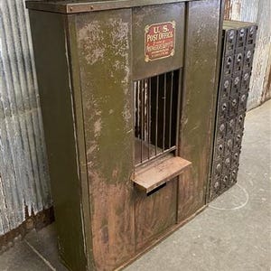 May include: An antique U.S. Post Office box with a weathered, olive green finish. The box features a service window with a metal grate, a small shelf, and a section of individual mailboxes. The U.S. Post Office logo is visible.