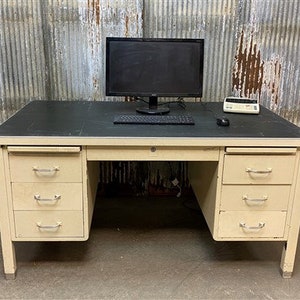 May include: A vintage metal desk with a black top and cream-colored drawers. The desk has three drawers on each side with silver handles. A computer monitor, keyboard, mouse, and a small electronic device sit on top.