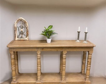May include: A light brown wooden console table with three sets of turned legs and a lower shelf. The table is in a room with white walls and a decorative windowpane hanging on the wall.