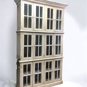 May include: A tall, white, wooden bookcase with glass doors and shelves. The bookcase has a distressed finish and is made of multiple sections.