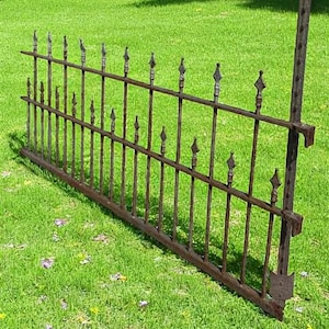 May include: A section of a weathered, wrought iron fence with decorative spear-shaped finials. The fence is a dark, rusty brown color and is set against a backdrop of green grass. The fence appears to be vintage or antique.