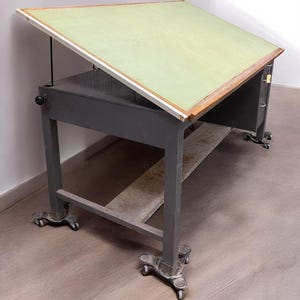 May include: An adjustable drafting table with a light green angled surface and a wooden border. The table has a gray metal frame, a lower shelf, and is mounted on wheels. The table's dimensions are approximately 36 inches wide and 48 inches long.