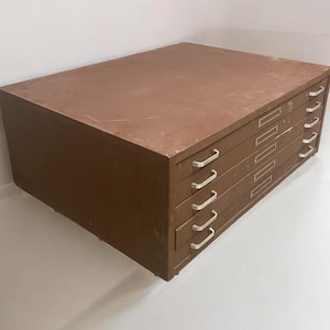 May include: A brown, vintage flat file cabinet with five drawers. Each drawer has a silver-colored handle and a label holder. The cabinet is rectangular and appears to be made of metal.