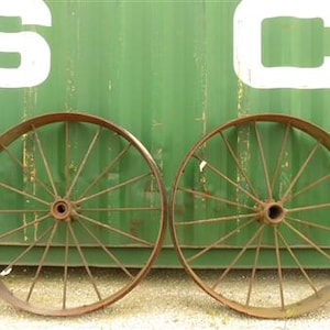 May include: Two antique wagon wheels with rusty brown metal rims and spokes. The wheels are set against a green background with large white letters. The wheels are circular and have a central hub.
