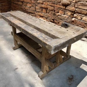 May include: A weathered, wooden workbench with a rectangular top and sturdy legs. The workbench features a central trough and is constructed from aged, gray-brown wood. The design suggests a vintage or antique aesthetic, suitable for a workshop or rustic decor.