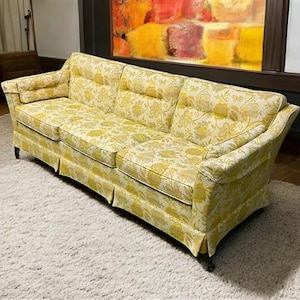 May include: A three-cushion sofa with a yellow floral pattern. The sofa has a skirted base and rolled arms. The fabric is a light yellow with a repeating floral design in a darker yellow. The sofa sits on short, dark legs.