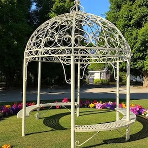 May include: A cream-colored, ornate metal gazebo with a domed roof and curved benches. The gazebo is surrounded by a bed of colorful flowers and green grass. The structure is in a park setting.