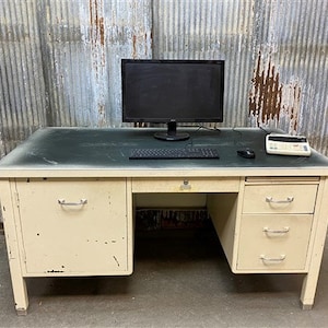 May include: A vintage metal desk with a light beige finish and a green laminate top. The desk has three drawers on the right side and one large drawer on the left side. A computer monitor, keyboard, mouse, and a small calculator are on the desk.