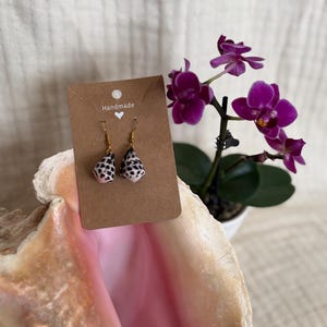 May include: A pair of dangle earrings featuring small, cone-shaped seashells with black spots on a light background. The earrings are attached to a rectangular brown card labeled with the word Handmade and a white heart icon.