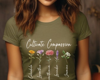 Puede incluir: Una camiseta verde con el texto "Cultivate Compassion" en escritura blanca. Cinco flores diferentes están ilustradas debajo del texto con sus nombres escritos verticalmente debajo de ellas: Calendula, Lisianthus, Crisantemo, Lavanda y Jazmín.