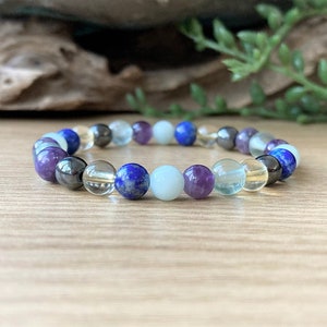 May include: A multi-gemstone bracelet with purple, blue, clear, and yellow beads. The bracelet is displayed on a light brown wooden surface. The beads are arranged in a circular pattern.