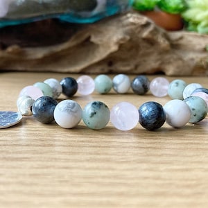 Puede incluir: Un brazalete hecho con cuentas blancas, verdes y negras con un dije de plata. El brazalete est&aacute; sobre una superficie de madera.