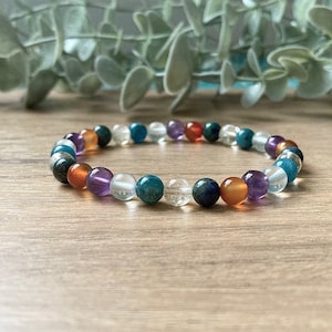 May include: A multi-coloured gemstone bracelet with beads in shades of blue, purple, orange, and clear. The bracelet is elasticated and is displayed on a wooden surface.