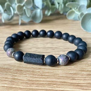 May include: A black beaded bracelet featuring a rectangular black stone and two speckled beads. The bracelet is made of matte black beads and is displayed on a wooden surface.