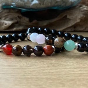 May include: Two bracelets with black beads and colourful gemstone beads. The top bracelet has a light green, brown, and red bead. The bottom bracelet has a white, brown, and red bead.