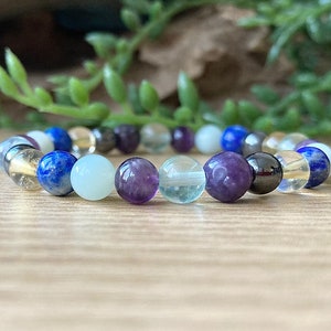 May include: A beaded bracelet featuring a variety of colorful gemstones. The bracelet includes blue lapis lazuli, clear quartz, purple amethyst, and other stones in shades of white, yellow, and black. The bracelet is strung on an elastic band.