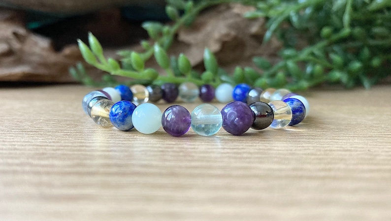 May include: A beaded bracelet featuring a variety of colorful gemstones. The bracelet includes blue lapis lazuli, clear quartz, purple amethyst, and other stones in shades of white, yellow, and black. The bracelet is strung on an elastic band.