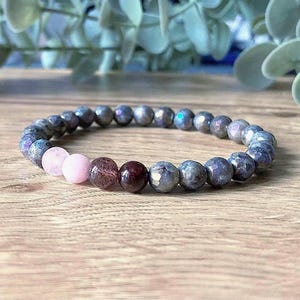 May include: A beaded bracelet featuring faceted grey labradorite beads with iridescent flashes, two smooth light pink beads, and two dark reddish-brown beads. The circular elastic bracelet is constructed from polished round stones arranged in a continuous loop.