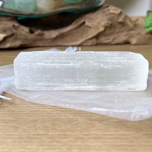 Pu&ograve; includere: Un cristallo di selenite bianco traslucido e rettangolare &egrave; appoggiato su un sacchetto di organza bianco trasparente. Il cristallo ha una consistenza a strati. Lo sfondo comprende una superficie in legno e vegetazione.