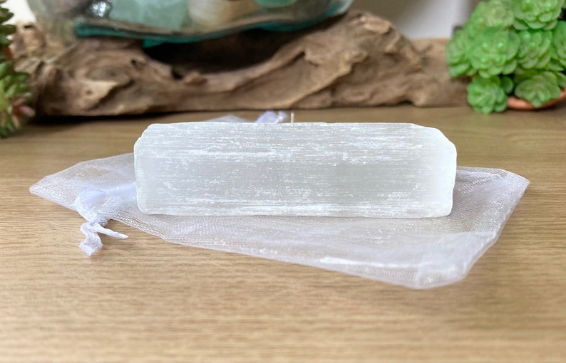 Pu&ograve; includere: Un cristallo di selenite bianco traslucido e rettangolare &egrave; appoggiato su un sacchetto di organza bianco trasparente. Il cristallo ha una consistenza a strati. Lo sfondo comprende una superficie in legno e vegetazione.