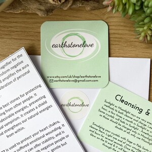 Pu&ograve; includere: Un insieme di biglietti stampati con il testo "earthstonelove" e informazioni sulla pulizia dei cristalli e la protezione energetica. I biglietti sono verde chiaro e bianchi, con uno sfondo in legno.