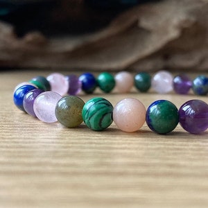 Peut inclure: Un bracelet compos&eacute; de perles de diff&eacute;rentes pierres pr&eacute;cieuses, dont du rose, du violet, du vert et du bleu. Les perles sont rondes et lisses, et le bracelet est enfil&eacute; sur un cordon &eacute;lastique transparent.