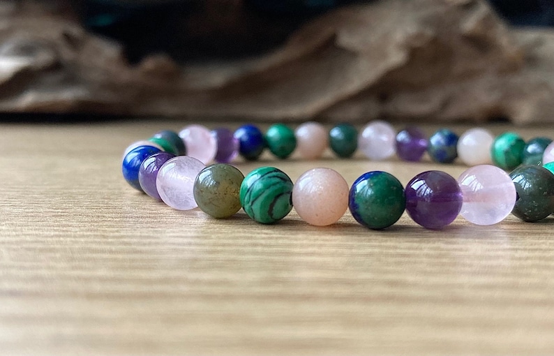Peut inclure: Un bracelet compos&eacute; de perles de diff&eacute;rentes pierres pr&eacute;cieuses, dont du rose, du violet, du vert et du bleu. Les perles sont rondes et lisses, et le bracelet est enfil&eacute; sur un cordon &eacute;lastique transparent.