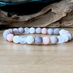 May include: A bracelet made of pink, gray, and white beads. The beads are round and have a smooth, polished finish. The bracelet is strung on a clear elastic cord.