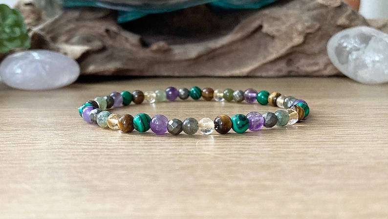 Pu&ograve; includere: Un braccialetto di perline con una variet&agrave; di pietre preziose nei toni del viola, verde, oro e trasparente. Il braccialetto &egrave; infilato su un cordino elastico e ha una circonferenza di circa 18 cm. Le pietre preziose includono ametista, malachite e occhio di tigre.