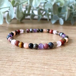 May include: A multi-coloured gemstone bracelet. The bracelet features beads in shades of red, pink, purple, brown, black, and clear. The bracelet is displayed on a wooden surface with green foliage in the background.