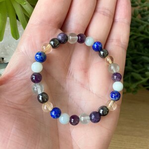 May include: A beaded bracelet featuring a variety of colorful gemstones. The bracelet includes round beads in shades of blue, purple, clear, yellow, and black. The bracelet is strung on an elastic band, and is shown on a person's hand.
