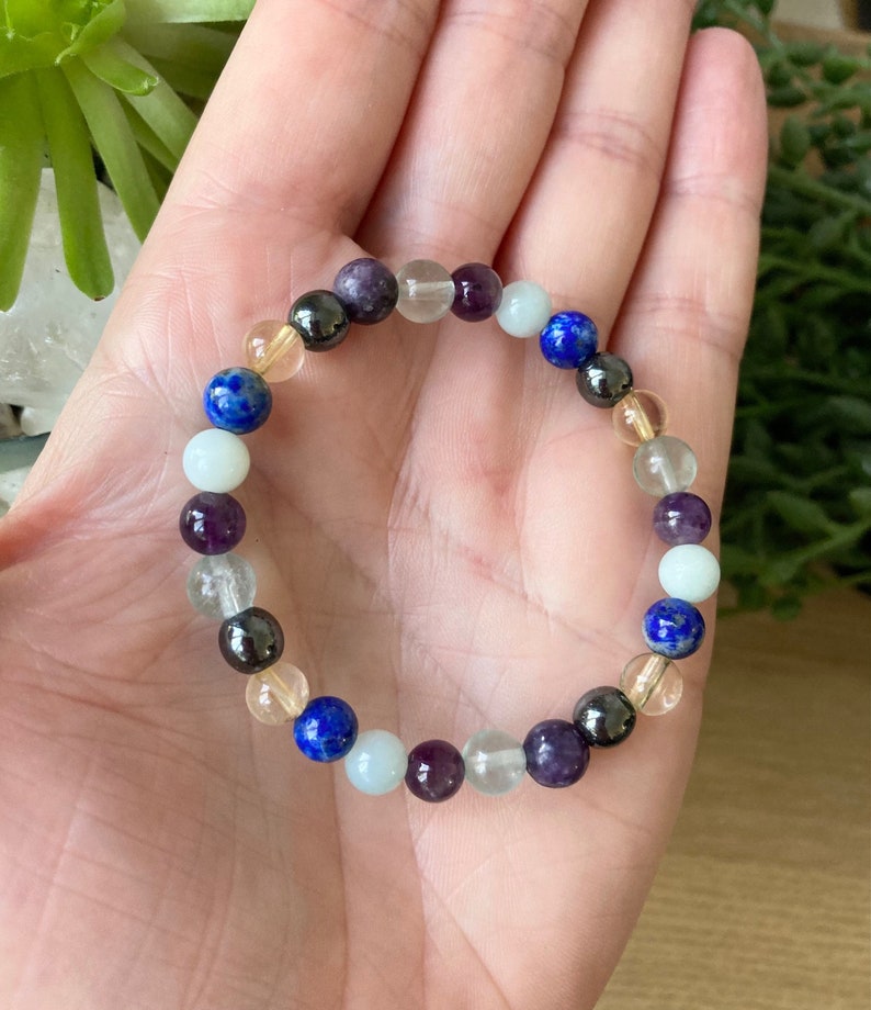 May include: A beaded bracelet featuring a variety of colorful gemstones. The bracelet includes round beads in shades of blue, purple, clear, yellow, and black. The bracelet is strung on an elastic band, and is shown on a person's hand.