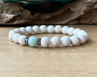 May include: White beaded bracelet with a light blue bead and silver accents. The bracelet features round, white beads and a central light blue bead, framed by silver-toned spacers with small, sparkling details. The bracelet is displayed on a wooden surface.