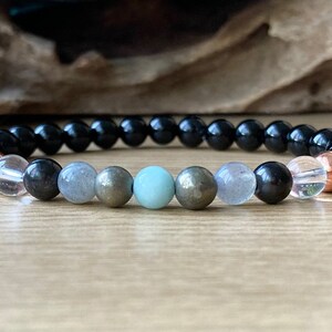 May include: A black beaded bracelet with a variety of gemstone beads, including clear, grey, and green beads. The bracelet also features two copper beads.