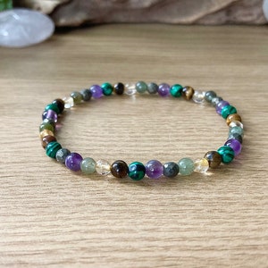 Pu&ograve; includere: Un braccialetto di perline con una variet&agrave; di pietre preziose colorate. Il braccialetto include perline verdi, viola, marroni e trasparenti. Il braccialetto &egrave; esposto su una superficie di legno.
