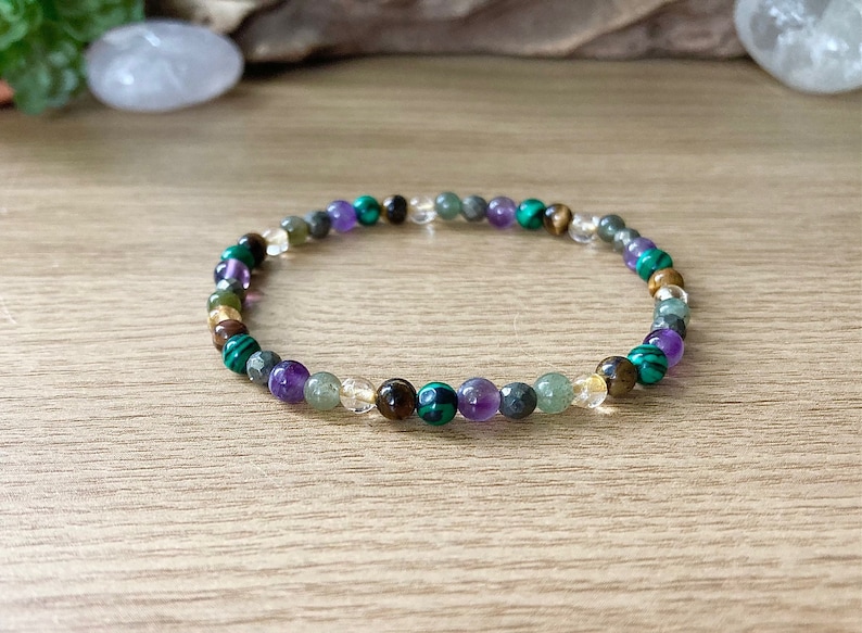Pu&ograve; includere: Un braccialetto di perline con una variet&agrave; di pietre preziose colorate. Il braccialetto include perline verdi, viola, marroni e trasparenti. Il braccialetto &egrave; esposto su una superficie di legno.