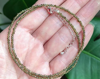 May include: A fine necklace composed of tiny, olive-green beads. The necklace has a silver-coloured clasp and an adjustable chain. The beads are evenly sized, creating a subtle, elegant appearance. The necklace is displayed against a soft background.