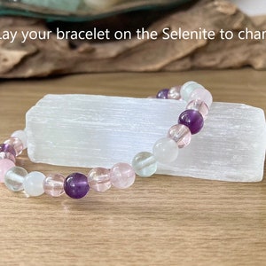 Pu&ograve; includere: Un braccialetto di perline con ametista, quarzo rosa e perline trasparenti poggia su una piastra di ricarica in selenite bianca. Il braccialetto &egrave; un mix di colori rosa, viola e trasparenti. Il testo nell'immagine recita: "Lay your bracelet on the Selenite to charge."