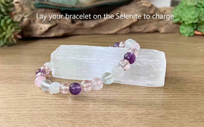 Pu&ograve; includere: Un braccialetto di perline con ametista, quarzo rosa e perline trasparenti poggia su una piastra di ricarica in selenite bianca. Il braccialetto &egrave; un mix di colori rosa, viola e trasparenti. Il testo nell'immagine recita: "Lay your bracelet on the Selenite to charge."