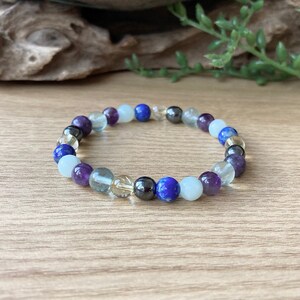 May include: A beaded bracelet featuring a variety of colorful gemstones. The bracelet includes purple, blue, clear, and black beads. The bracelet is displayed on a wooden surface.
