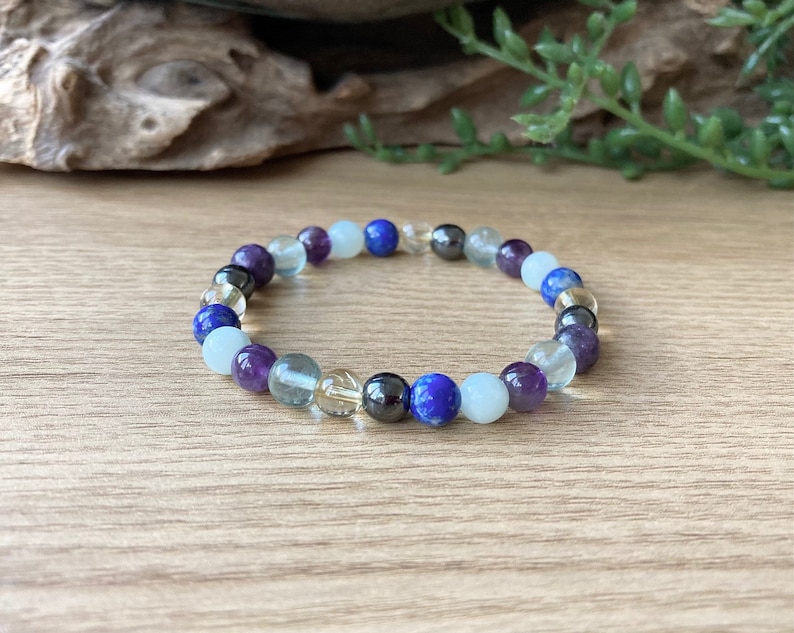 May include: A beaded bracelet featuring a variety of colorful gemstones. The bracelet includes purple, blue, clear, and black beads. The bracelet is displayed on a wooden surface.