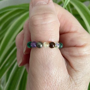 May include: A ring made of various coloured gemstones, including green, purple, brown, gold, and clear. The ring is worn on a person's finger.