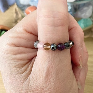 May include: A bracelet made of small, round, colourful beads. The beads are in a variety of colours, including pink, brown, grey, black, and purple. The bracelet is worn on a person's wrist.