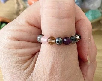 May include: A bracelet made of small, round, colorful beads. The beads are in a variety of colors, including pink, brown, gray, black, and purple. The bracelet is worn on a person's wrist.