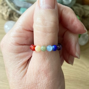 May include: A rainbow chakra bracelet made with round, colourful gemstone beads. The bracelet is worn on a person's thumb.