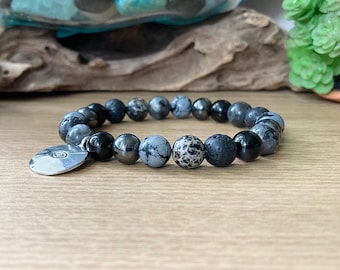 May include: A black, gray, and silver beaded bracelet with a round silver charm. The bracelet is made with lava rock, black onyx, and snowflake obsidian beads.
