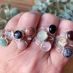 May include: A collection of silver rings featuring a variety of gemstone cabochons, including black, pink, green, white, and brown. The rings are stacked on a hand.