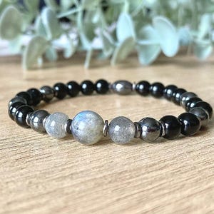 May include: A beaded bracelet featuring a mix of black, gray, and iridescent blue-gray beads. The bracelet is made of round beads and is displayed on a wooden surface. The bracelet is a piece of jewelry.