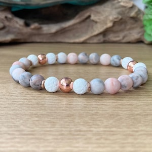 May include: A bracelet made of pink, gray, and white beads with rose gold accents. The bracelet is made of natural stones and is designed to be worn on the wrist.