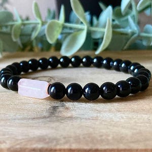 May include: A stretch bracelet composed of smooth, round, glossy black beads and a single, rectangular, pale pink stone bead. The beads are strung together in a circular pattern, creating a continuous loop of dark and light contrasting elements.